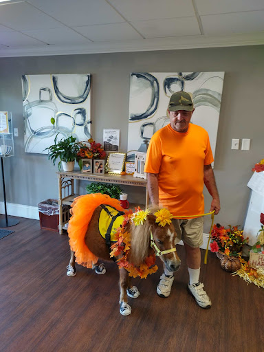 A man in an orange shirt stands next to a beautifully decorated miniature horse in a cozy indoor space.