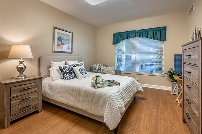 A cozy bedroom with a neatly made bed, wooden flooring, and a large window with green curtains.