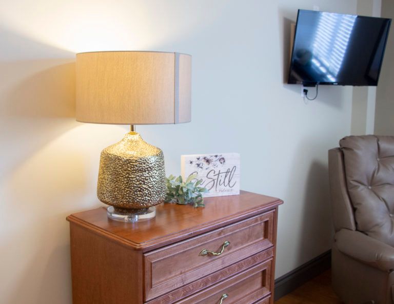 A cozy living room corner featuring a lamp on a wooden dresser with a decorative card and plant, alongside a wall-mounted TV.