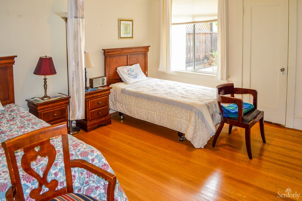 A cozy bedroom with a single bed and wooden furniture, featuring a nightstand, lamp, and a window letting in natural light.