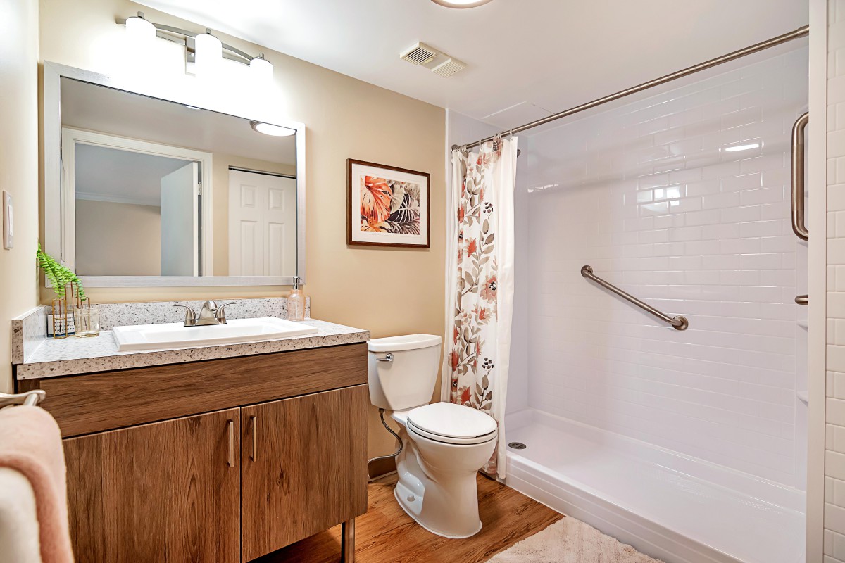 A modern bathroom with a wooden vanity, sink, toilet, and a shower with a grab bar.