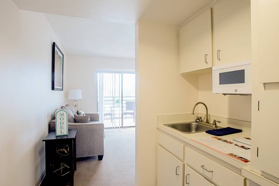 A small kitchen area with white cabinets and a sink, adjacent to a cozy living room with natural light.