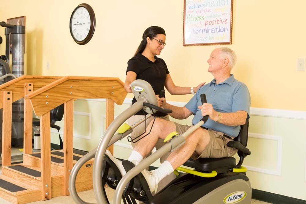 An elderly man receives guidance from a trainer while using an exercise machine for rehabilitation.