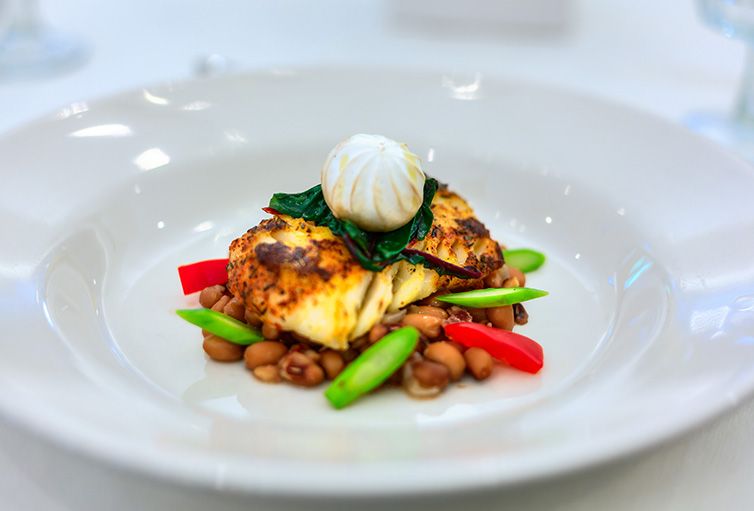 A gourmet dish featuring a grilled protein served with vibrant vegetables and beans, topped with a decorative garnish.