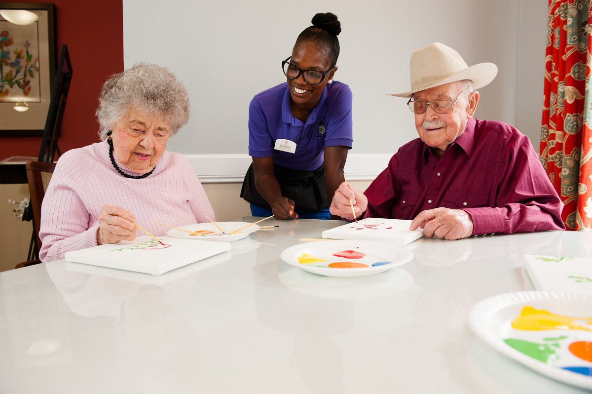 A caregiver assists elderly individuals during a painting activity at a seniors' home.