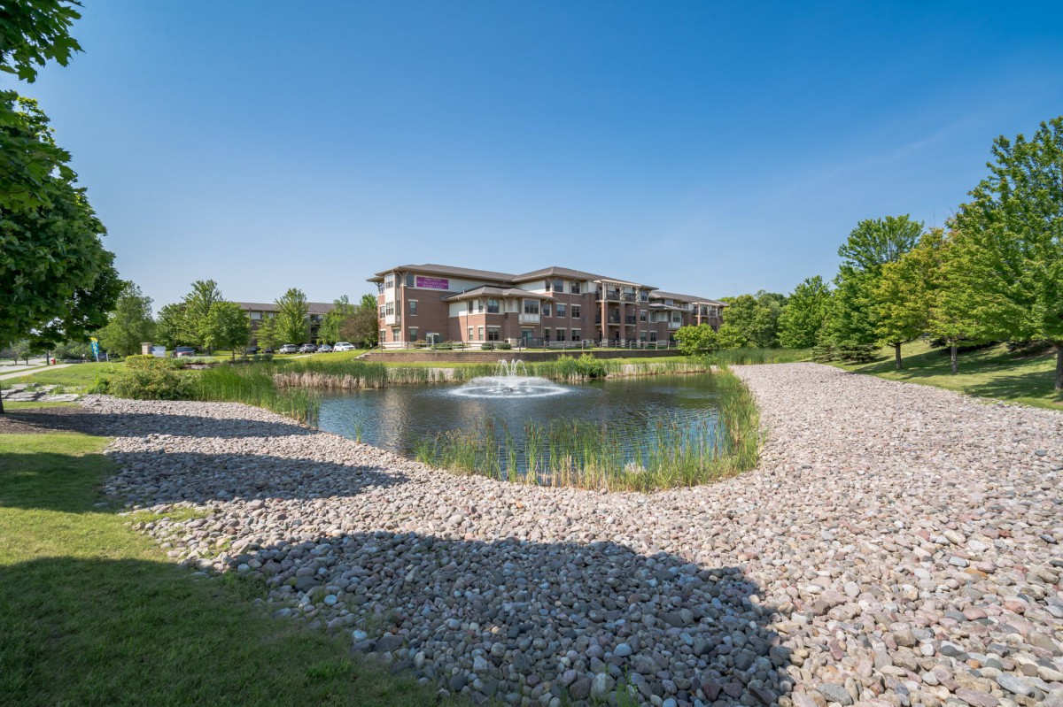Scenic view of a building by a pond with a fountain, surrounded by gravel paths and trees under a clear blue sky.
