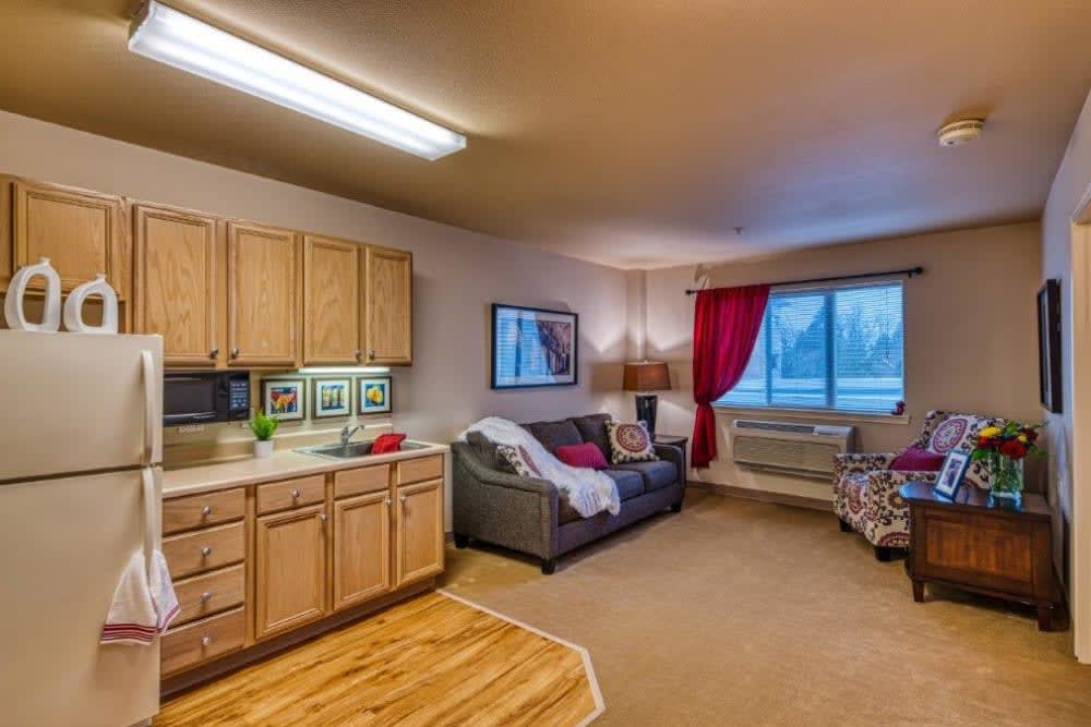 A cozy apartment with a small kitchenette and living area, featuring a sofa, window with red curtains, and wall art.
