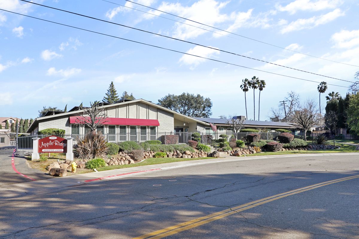A suburban building with red awnings, surrounded by a landscaped area and palm trees, next to a road under a clear blue sky.