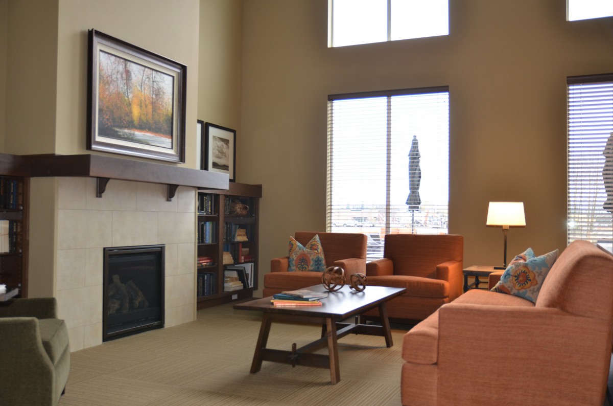 A cozy living room with a fireplace, orange armchairs, and a sofa centered around a wooden coffee table. Large windows provide natural light, enhancing the warm atmosphere.