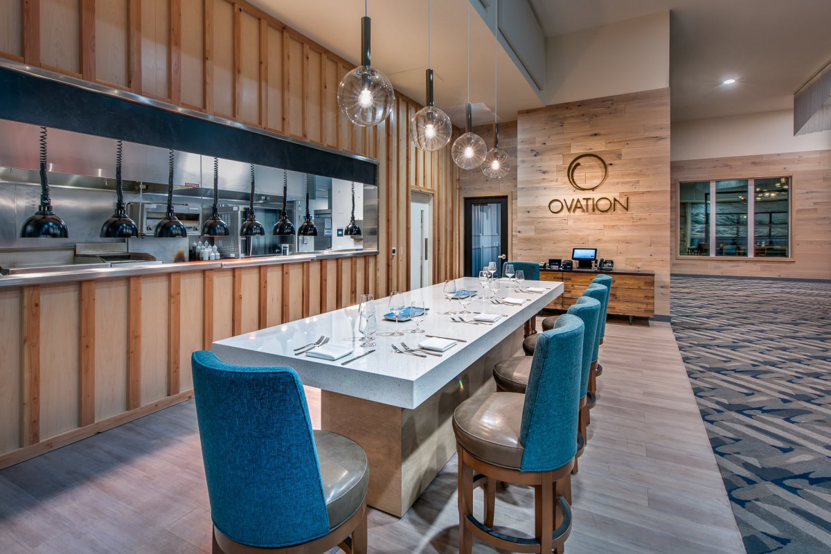 A modern restaurant interior featuring a long dining table with blue chairs and an open kitchen.