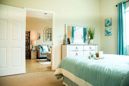 A cozy bedroom with a light and airy interior design, featuring a bed with teal bedding, a dresser with a mirror, and decorative plants.