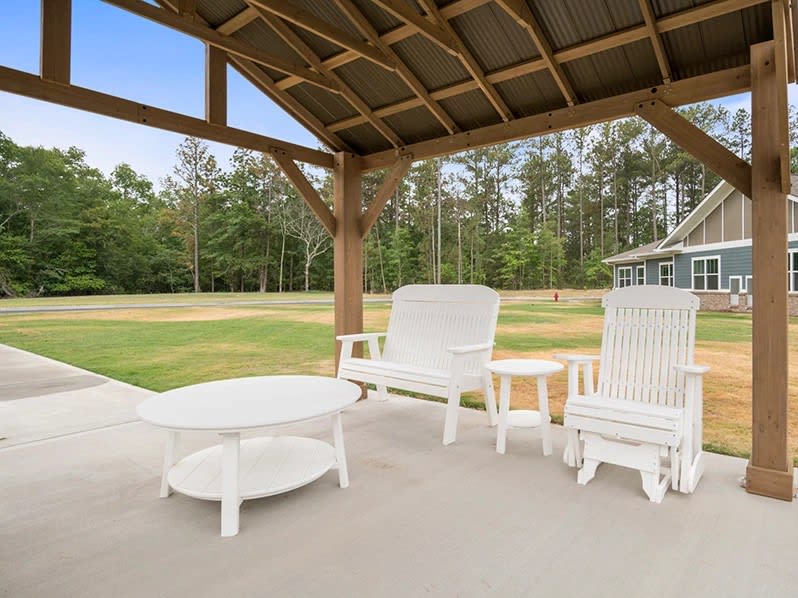 A cozy patio area with white furniture under a wooden pergola, overlooking lush greenery and trees.
