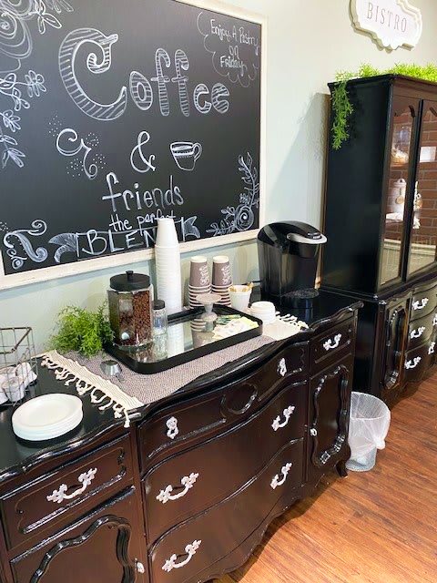 A cozy coffee station featuring a black chalkboard, coffee machine, cups, and decorative plants on dark furniture.