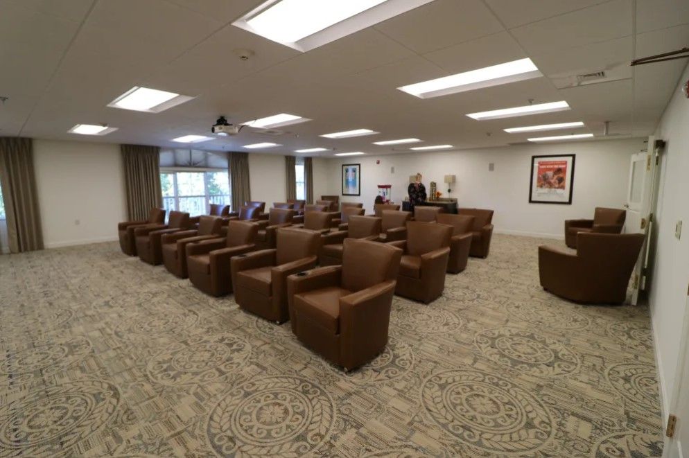 A spacious conference room with brown chairs arranged in rows, well-lit by overhead lights, and featuring large windows on one side.