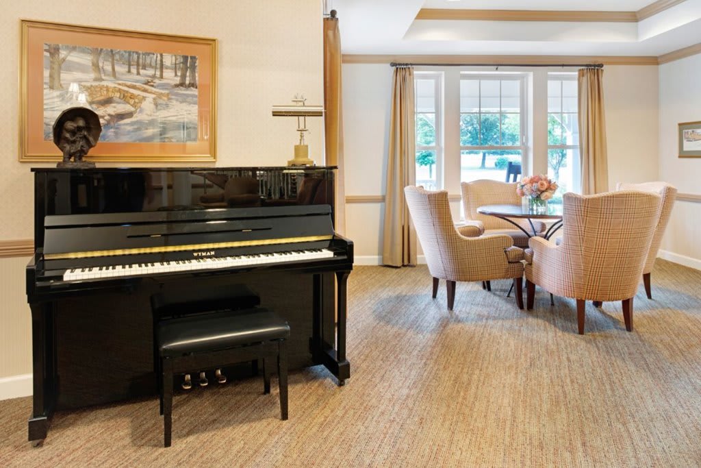 A cozy living space featuring a black upright piano and a dining area with chairs near a large window.