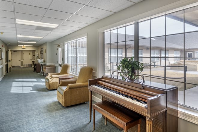 A cozy lounge area featuring a piano and comfortable chairs with ample natural light from large windows.