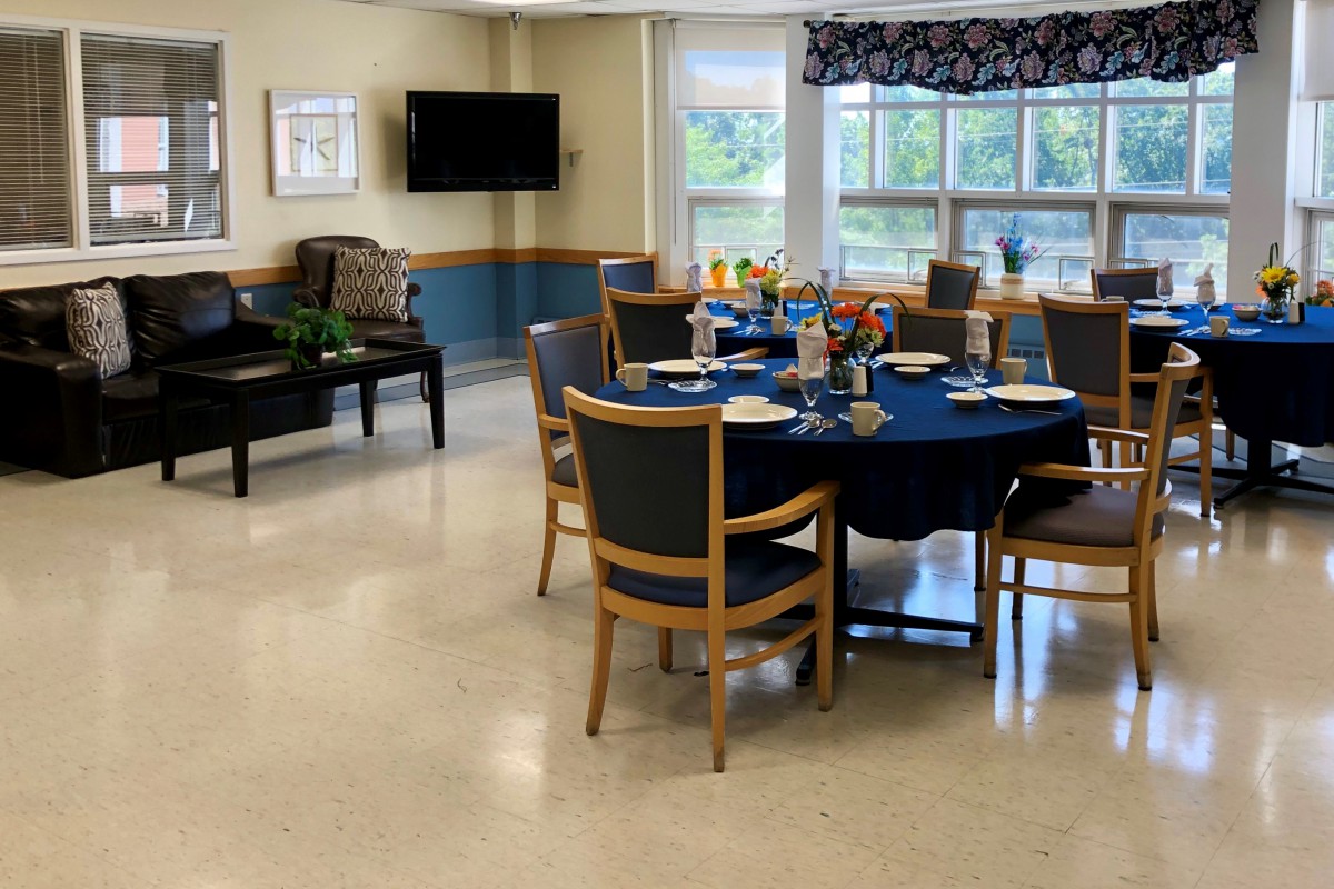A dining room with a round table set for a meal, featuring several chairs, a wall-mounted TV, and large windows letting in natural light.
