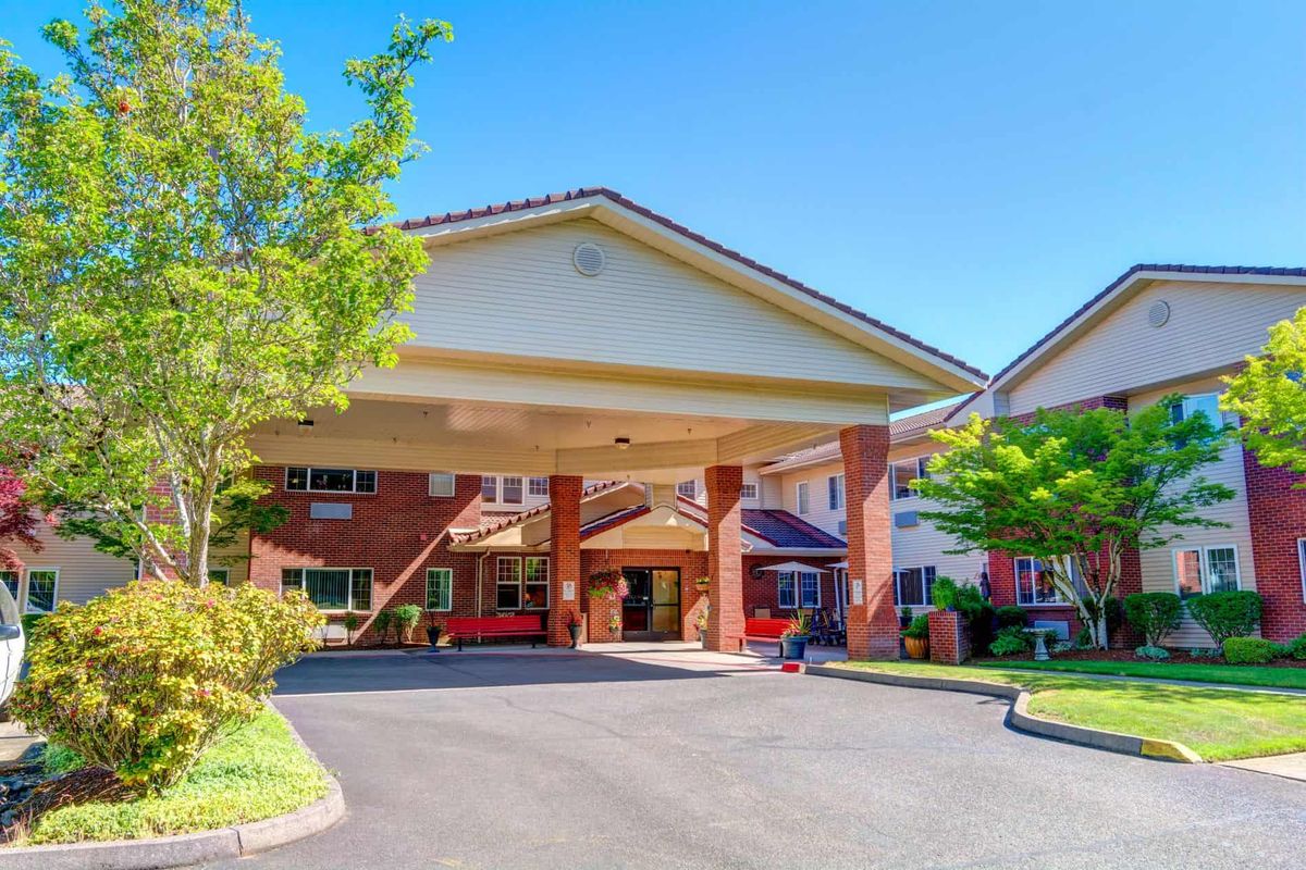A sunny day view of a building entrance with a covered driveway and surrounding greenery.