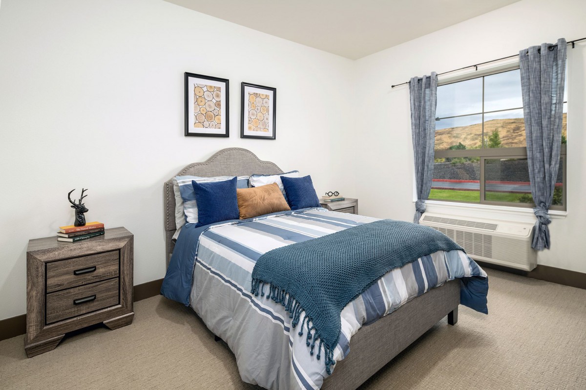 A cozy bedroom with a neatly made bed, featuring blue and gray bedding, flanked by nightstands and adorned with artwork above the headboard.