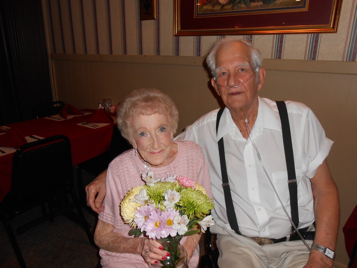 An elderly couple sitting together indoors, holding a bouquet of flowers.