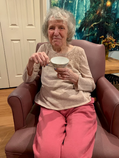A smiling elderly woman seated comfortably, enjoying a cup.