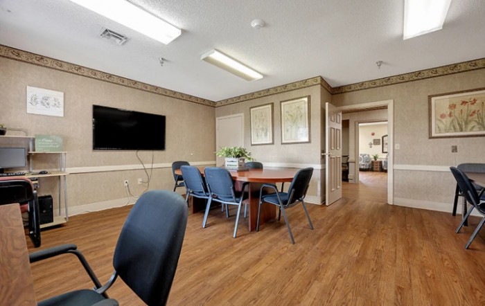 A well-lit conference room featuring a large table surrounded by chairs, a wall-mounted television, and wooden flooring.