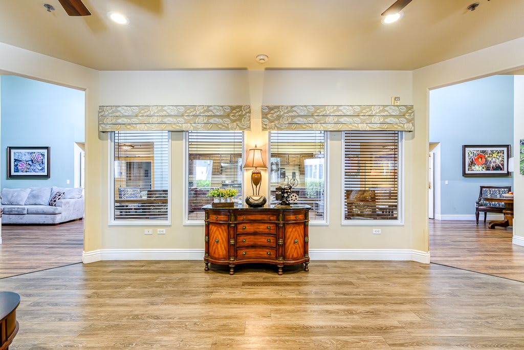 A well-lit living room interior featuring a wooden sideboard, lamp, and large windows.