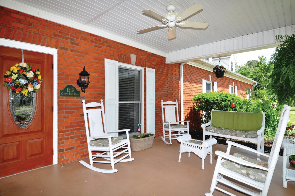 A cozy porch with white rocking chairs and a ceiling fan, surrounded by a red brick wall and greenery.