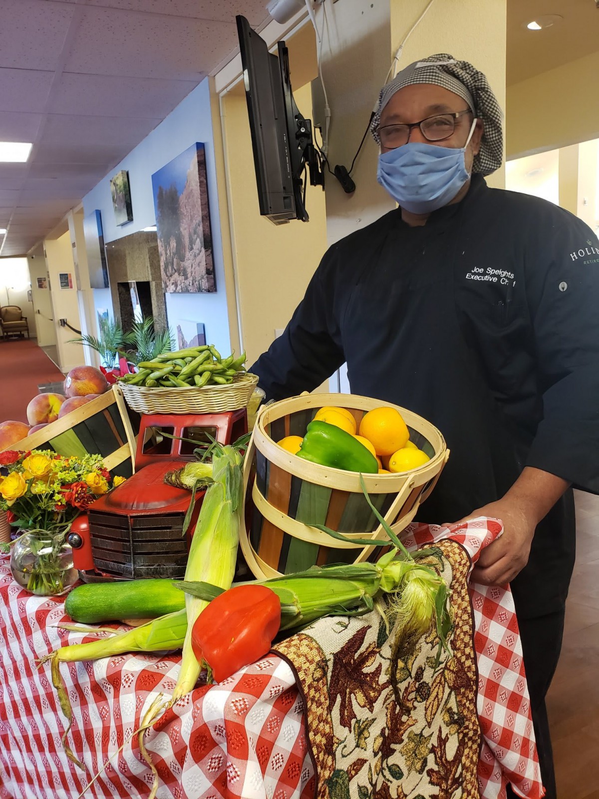 A chef wearing a mask stands next to a colorful display of vegetables and fruits in baskets.
