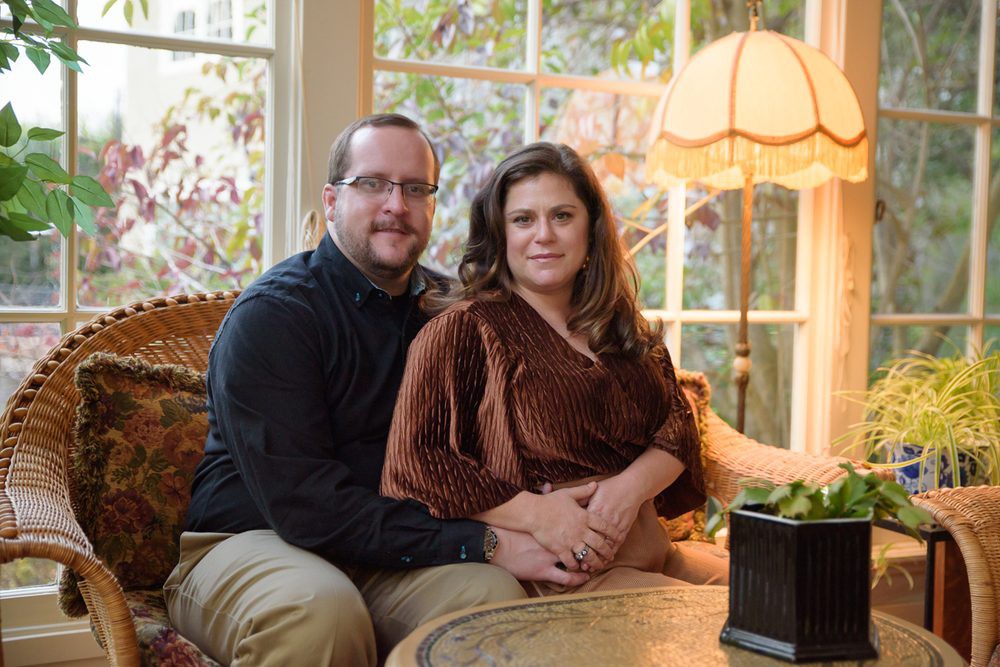 A couple sitting together on a sofa in a cozy living room with a large window and lamp.