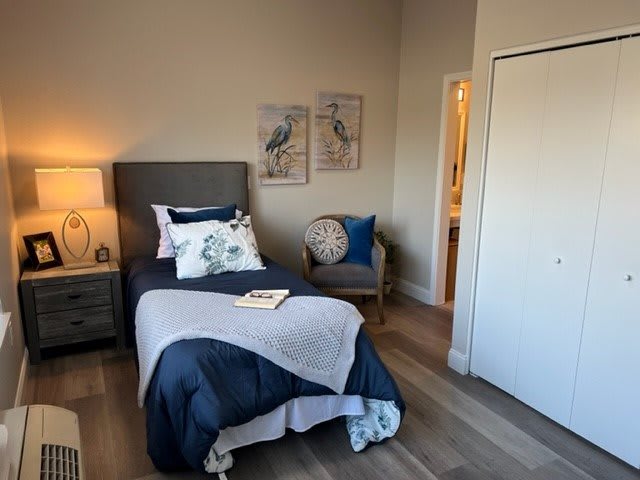 A cozy bedroom featuring a single bed with decorative pillows, a nightstand with a lamp, and minimalistic wall art.