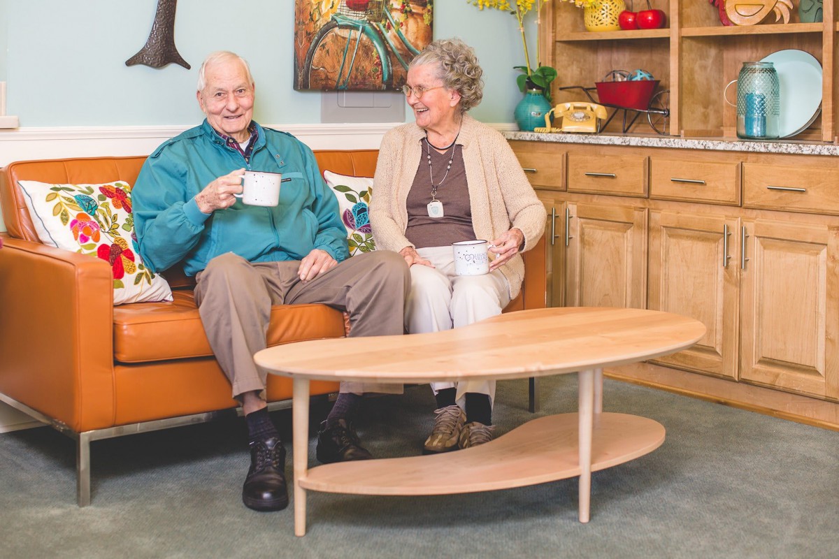 An elderly couple sitting on a sofa, smiling and enjoying coffee together in a cozy living room.