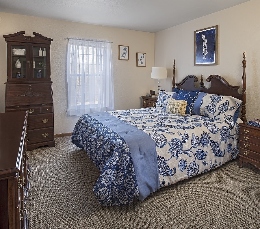 A cozy bedroom featuring a wooden bed with blue floral quilted bedding and matching pillows, accompanied by classic wooden furniture and illuminated by natural light from a window.