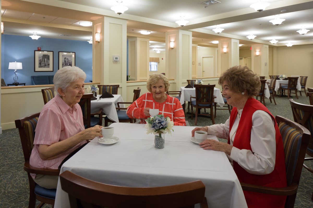 Three senior women enjoy a meal together, engaging in conversation at a dining facility.