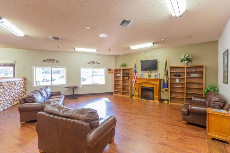 A spacious living room with comfortable seating, a fireplace, and a television mounted above. The room features bookshelves and flags, along with a wooden floor.