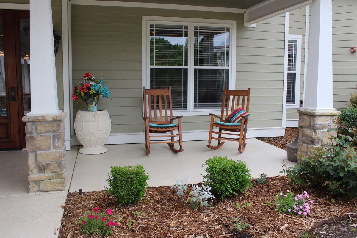 A cozy porch with two wooden rocking chairs and a decorative vase, surrounded by a small garden.