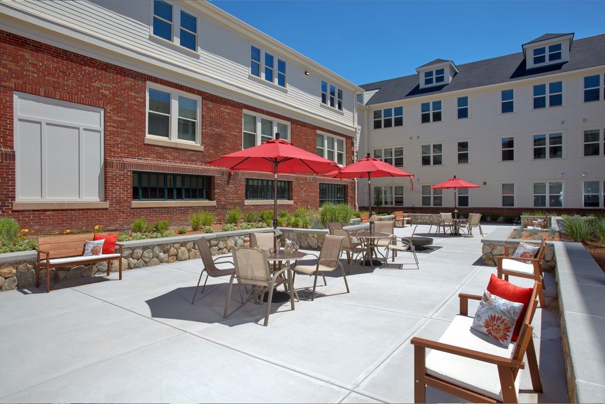 A sunny outdoor patio area with red umbrellas, seating, and a residential building in the background.