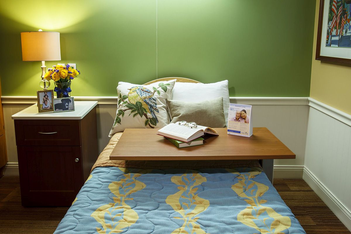 A cozy bedroom setup with a bed featuring decorative pillows, a bedside table with a lamp and flowers, and a book resting on a wooden tray.