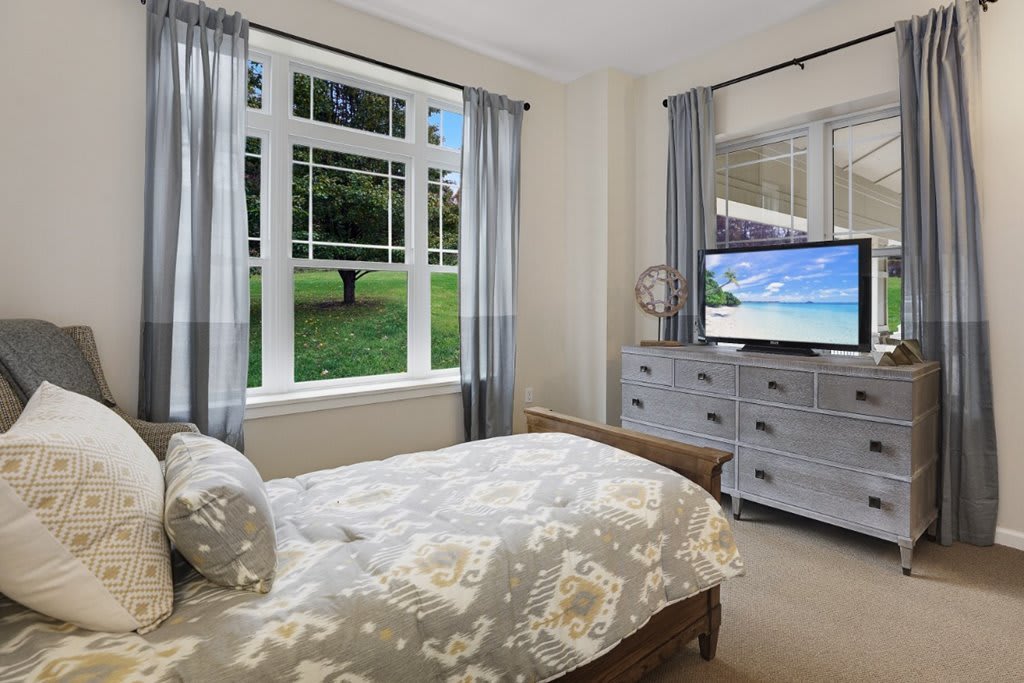 A cozy bedroom with a large window, curtains, and a television on a dresser.