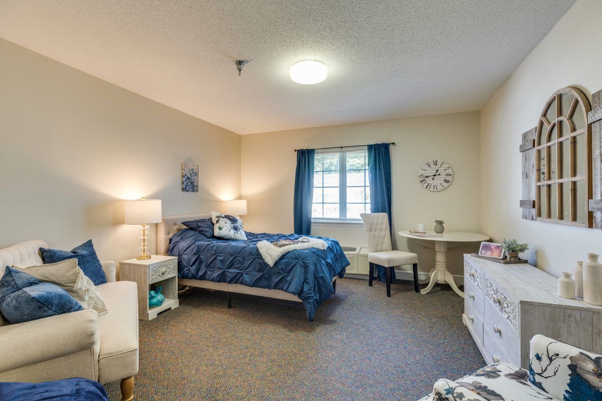A cozy bedroom with a bed, nightstand, chair, and table, featuring blue accents and natural lighting.