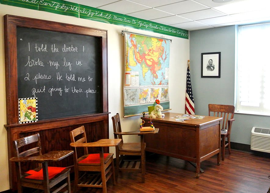 A vintage-style classroom with a chalkboard, wooden desk, and chairs, adorned with a world map and an American flag.