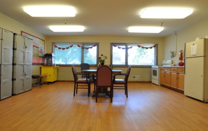 A well-lit dining room with a central table and chairs, featuring large windows and hardwood flooring.