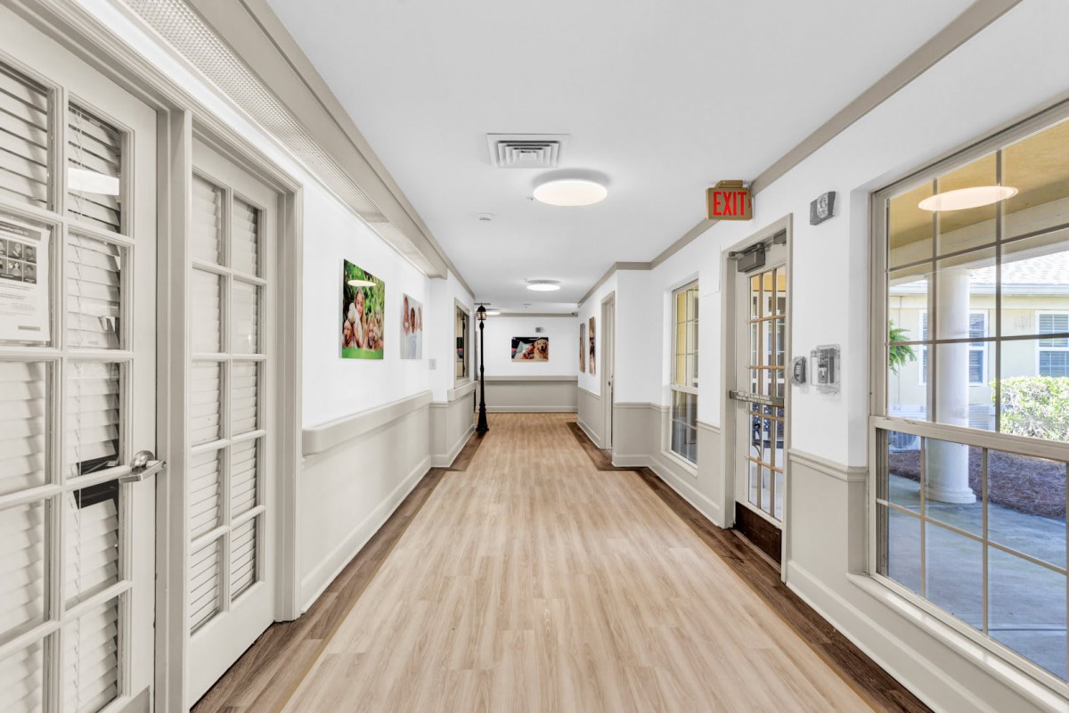 A clean and bright hallway in a healthcare facility with wooden floors, framed artwork on the walls, and closed doors.
