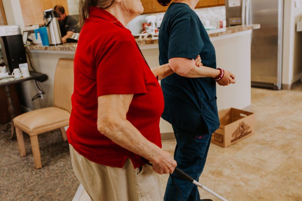 An elderly person with a walking stick receiving assistance from a caregiver indoors.