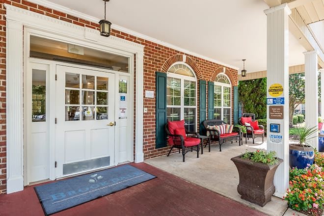 A welcoming front porch with red cushioned chairs and potted plants against a brick wall.
