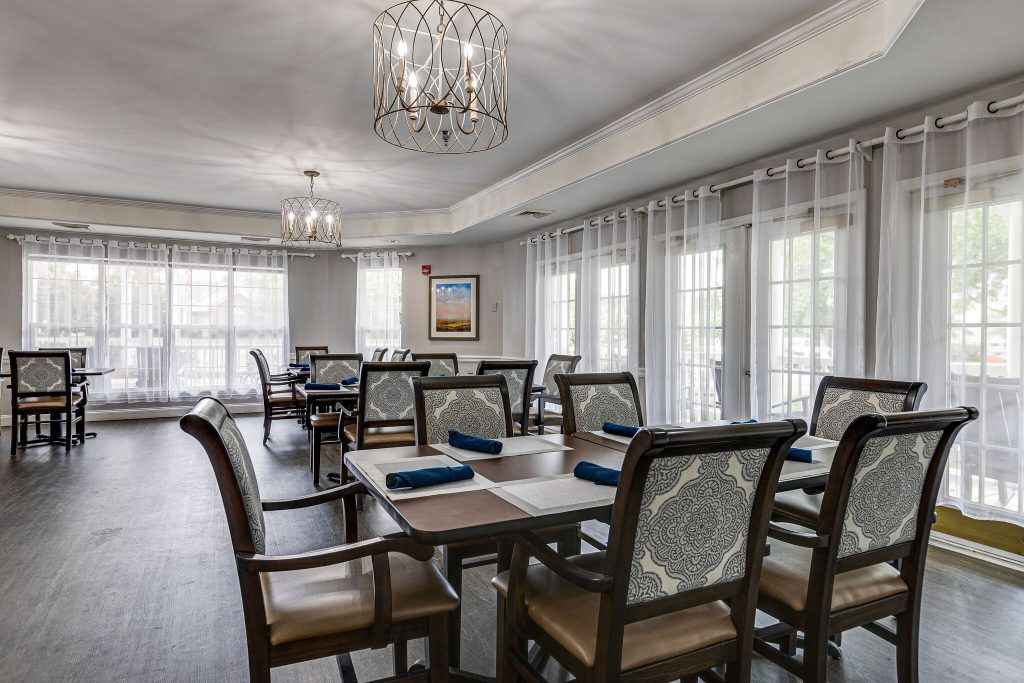 A well-lit dining room with neatly arranged tables and chairs, featuring elegant chandeliers and large windows with sheer curtains.