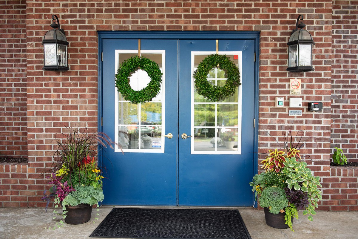A pair of blue double doors with green wreaths, surrounded by plants, set against a brick wall.