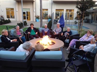 A group of seniors enjoys socializing around a fire pit in an outdoor seating area.