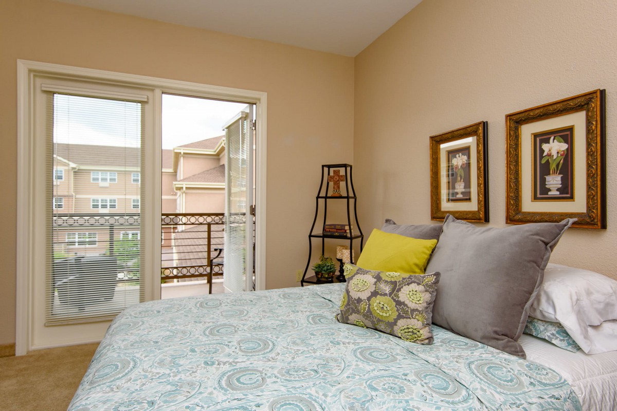 A cozy bedroom with elegant decor, featuring a neatly made bed with patterned bedding and colorful pillows, adjacent to a small balcony allowing natural light to brighten the space.