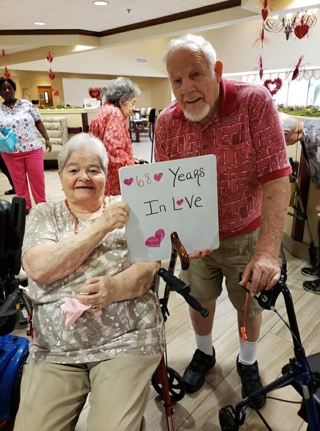 An elderly couple happily holding a sign that says 'Years In Love' during a community event.